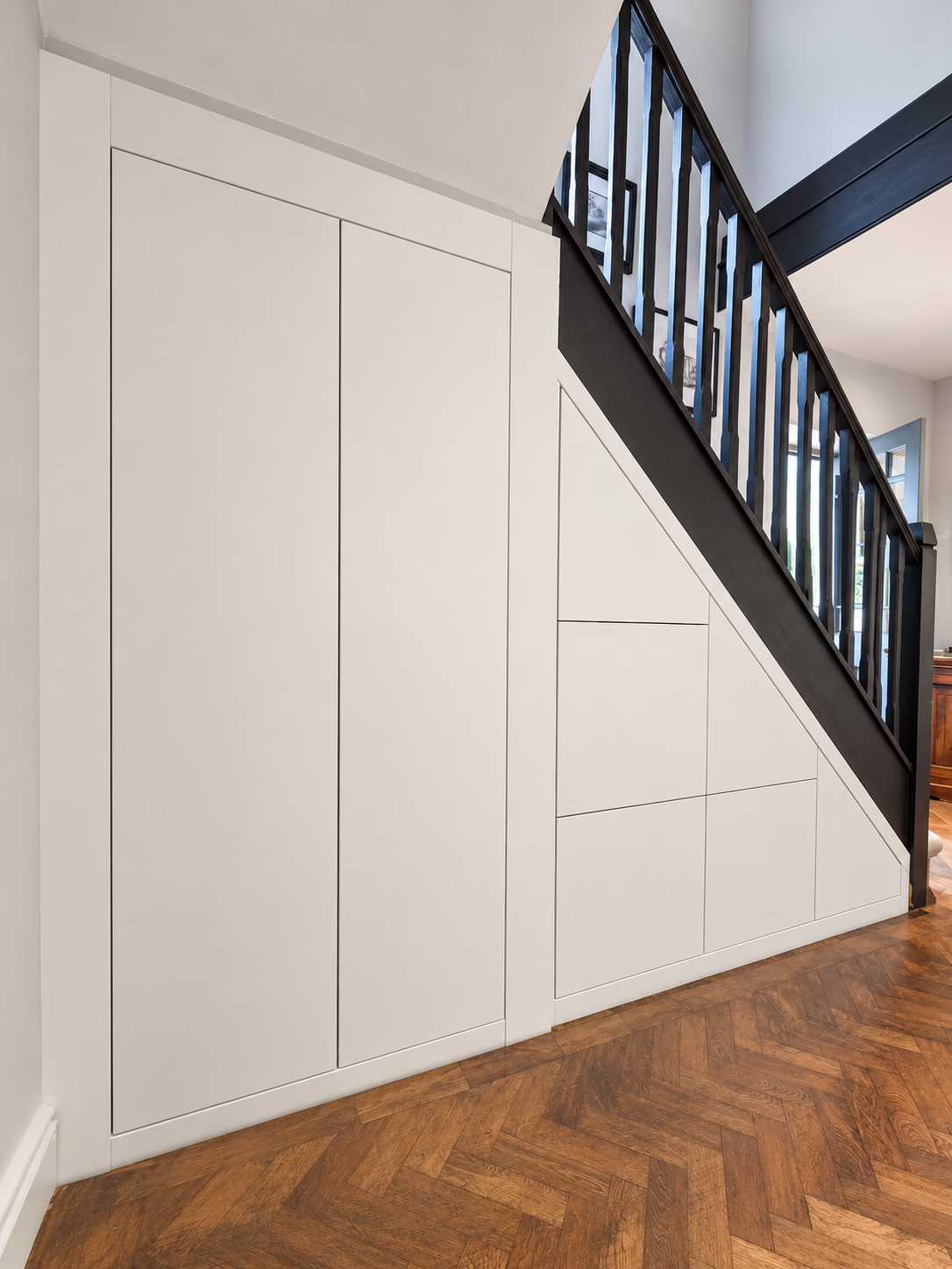 Handleless white under-stairs cupboards and drawers fitted beneath a painted staircase on herringbone flooring